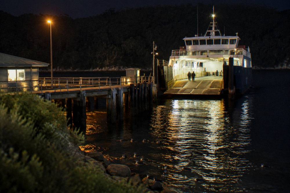 A vehicle ferry loading at a timber jetty with forested hills across the water