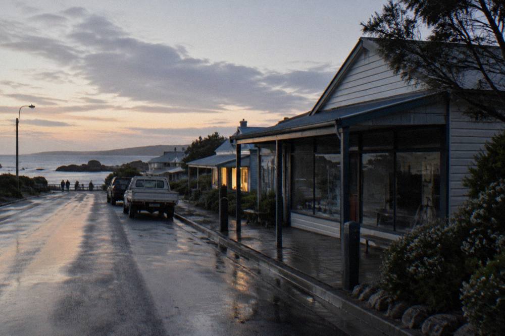 A quiet street in a small coastal town at dusk with a lit shopfront and parked cars