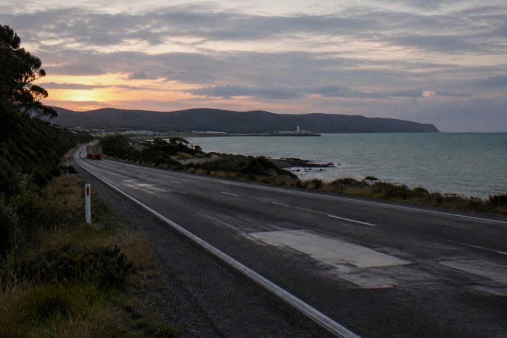 A coastal road at dusk with dark hills and a pale evening sky over the sea