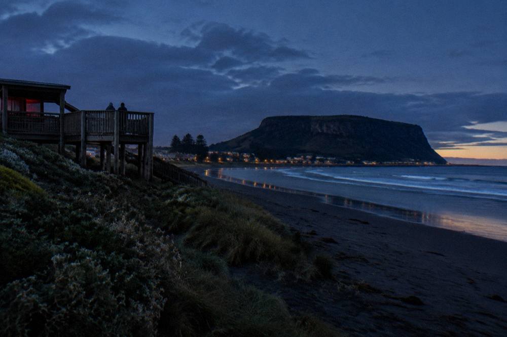 A wooden viewing platform above a dark beach at dusk with the lights of a small coastal town in the distance