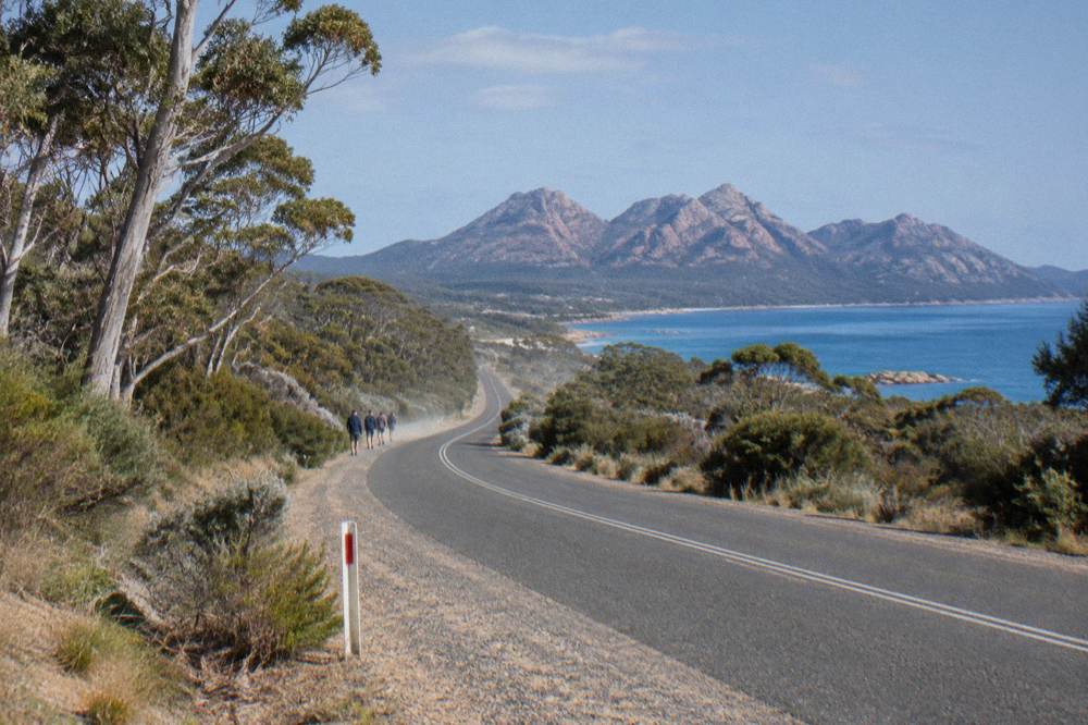 A sealed coastal highway curving toward pink granite mountains across blue Tasmanian sea