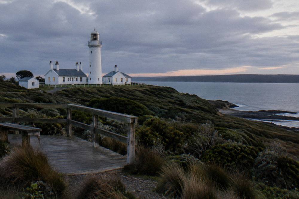 A small lighthouse above a dark coastal reserve with a viewing platform at dusk