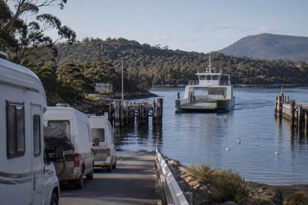 A vehicle ferry approaching a timber jetty on a calm forested channel