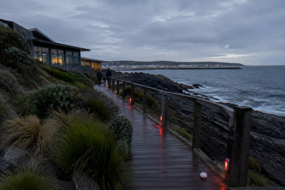 A timber boardwalk along a darkened beachfront with low red lighting near the rail